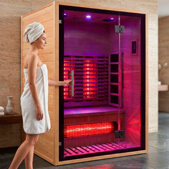 Woman in a towel with a towel wrapped on her head stands beside an open wooden infrared sauna glowing pink-red lights inside.