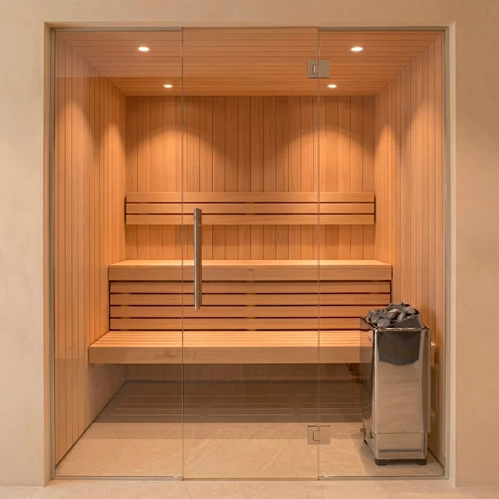 Glass-enclosed wooden sauna with two-tier benches and a metal heater on the right, stones on top, in a softly lit room.